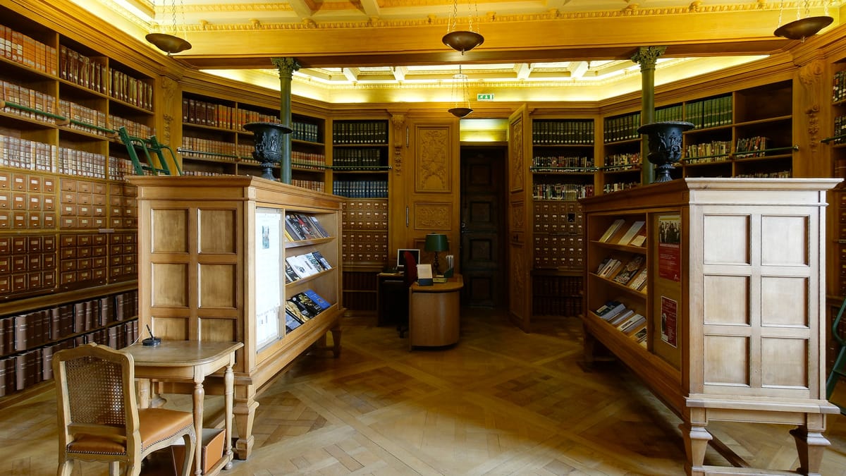 Salle de bibliothèque aux boiseries claires, avec des rayonnages de livres reliés, des meubles à tiroirs de fiches et des présentoirs, sans personne