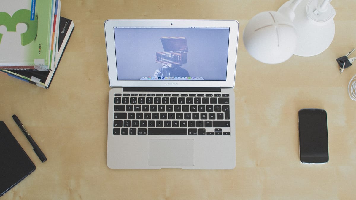 Vue de dessus d'un bureau en bois clair avec un ordinateur portable ouvert, un téléphone, des livres, un stylo et une lampe