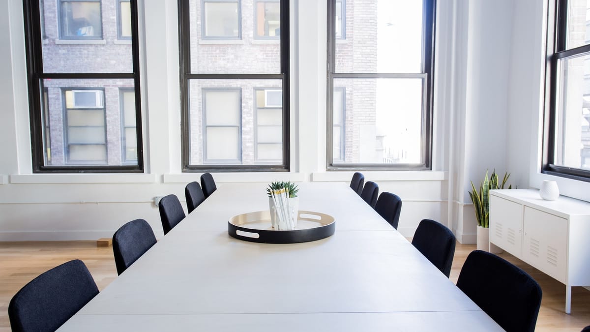 Salle de réunion vide et lumineuse : longue table blanche entourée de chaises noires, devant de grandes fenêtres