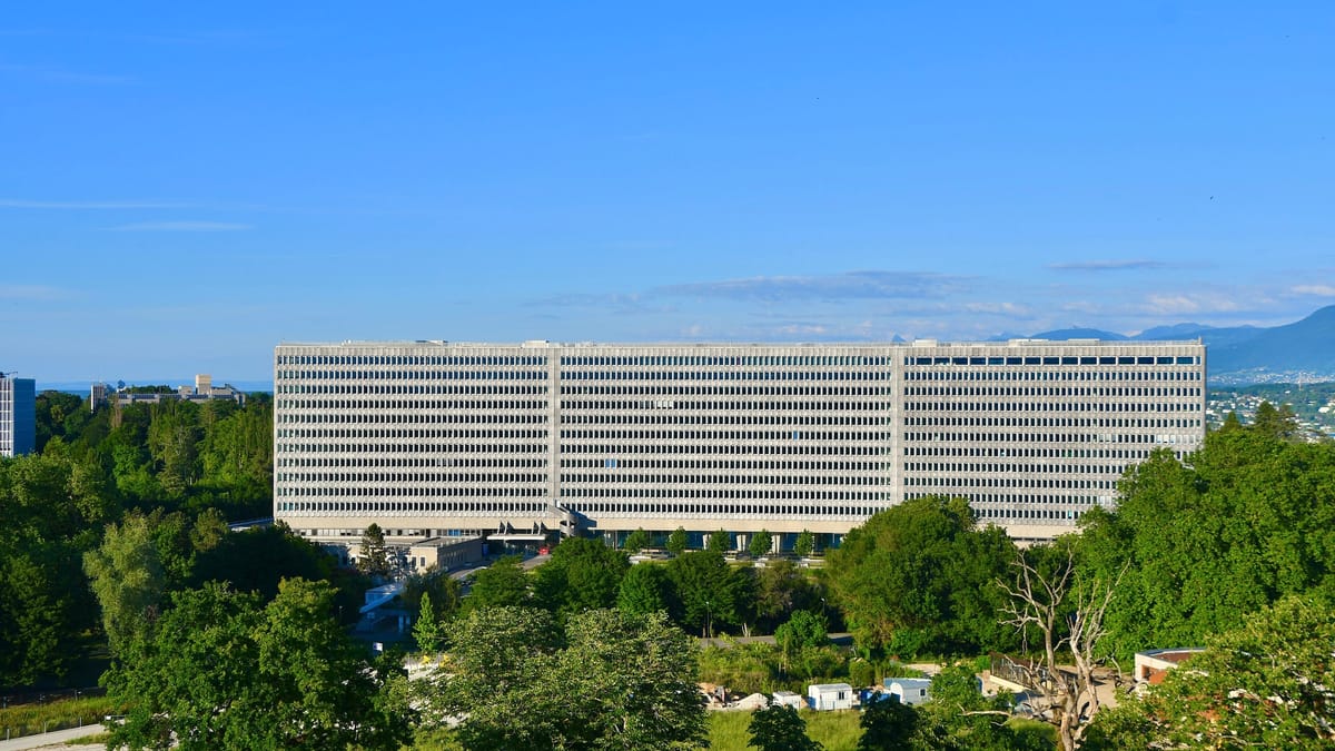 Long bâtiment de bureaux aux nombreuses fenêtres, entouré d’arbres, sous un ciel bleu, avec des montagnes à l’horizon